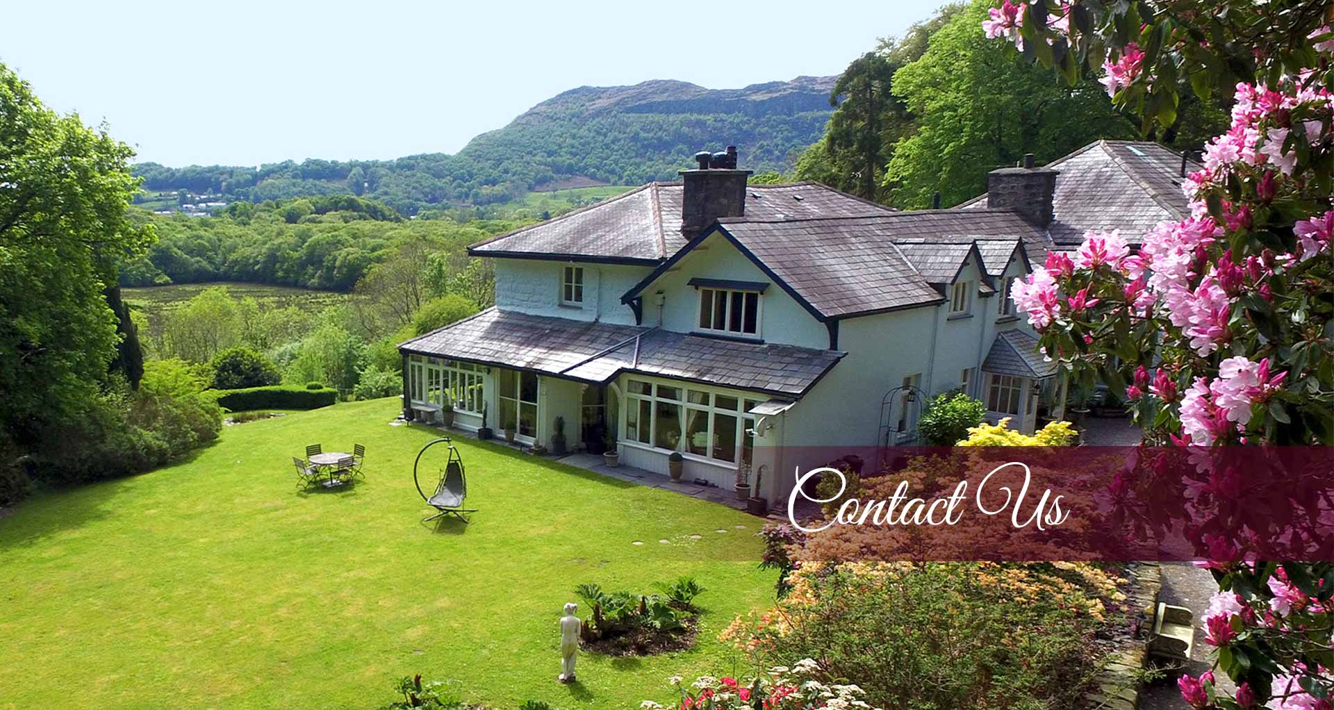 Contact Us banner showing aerial side elevation of the house taken in the Spring - Spring shrubs including a pink rhododendron in bloom on the right - Overlooking Shelleys lawn with panoramic view over Moel y Gest in the background - At Plas Tan-Yr-Allt - 5-Star luxury, literary & historic Country House B&B in Tremadog, Porthmadog, Snowdonia.